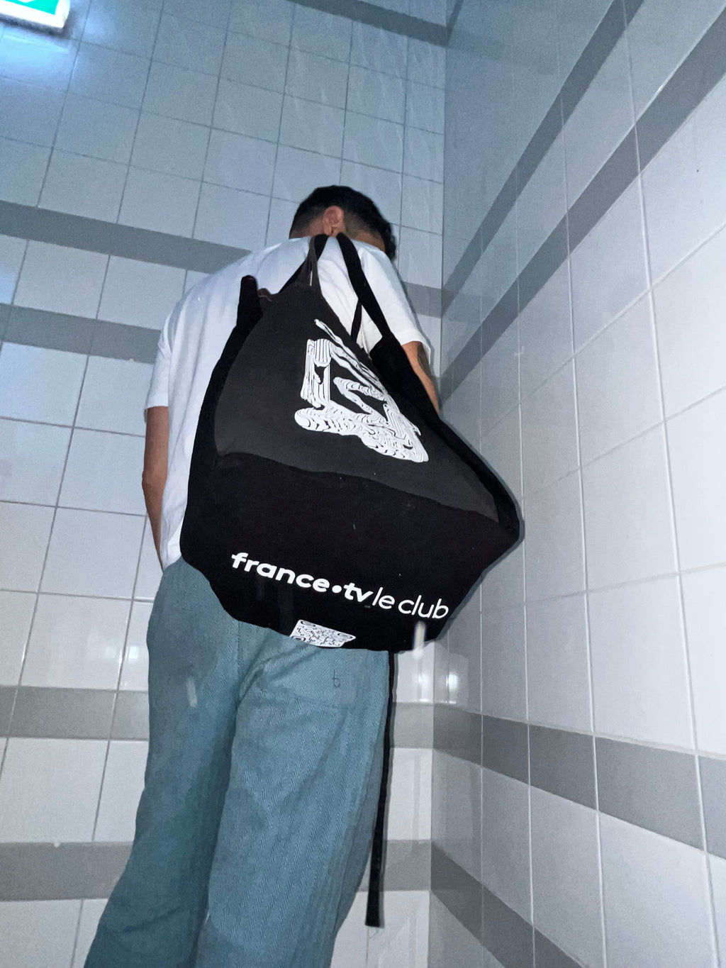 A person wearing a white t-shirt and blue jeans is shown from behind carrying a large black tote bag with white text reading 'france.tv.