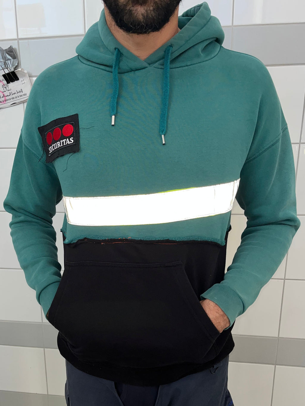 A man wearing a teal and black color-blocked hoodie with a reflective white horizontal stripe across the chest and a black patch on the left sleeve. The hoodie has a kangaroo pocket on the front.